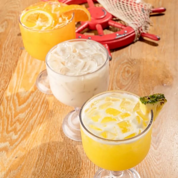 Three refreshing drinks, orange, horchata, and pineapple, on a wooden table with a decorative ship's wheel.