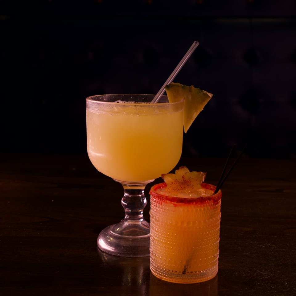 Two cocktails on a dark wooden table: a yellow margarita with pineapple and a spiced orange drink.