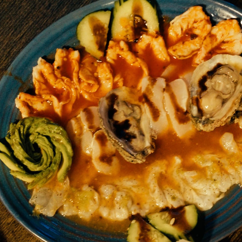 A blue plate of Mariscos La Riviera Nayarit seafood with shrimp, oysters, scallops, avocado, and cucumber.