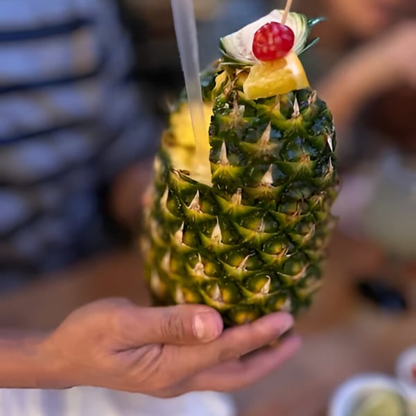 A person holds a hollowed-out pineapple filled with a drink, garnished with fruit and a straw.