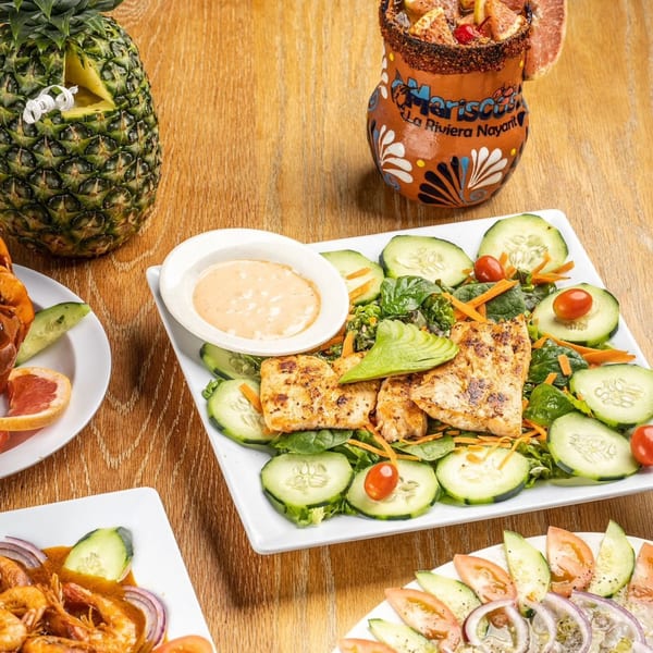 Grilled fish salad with avocado, cucumbers, and tomatoes, next to a pineapple and a drink.