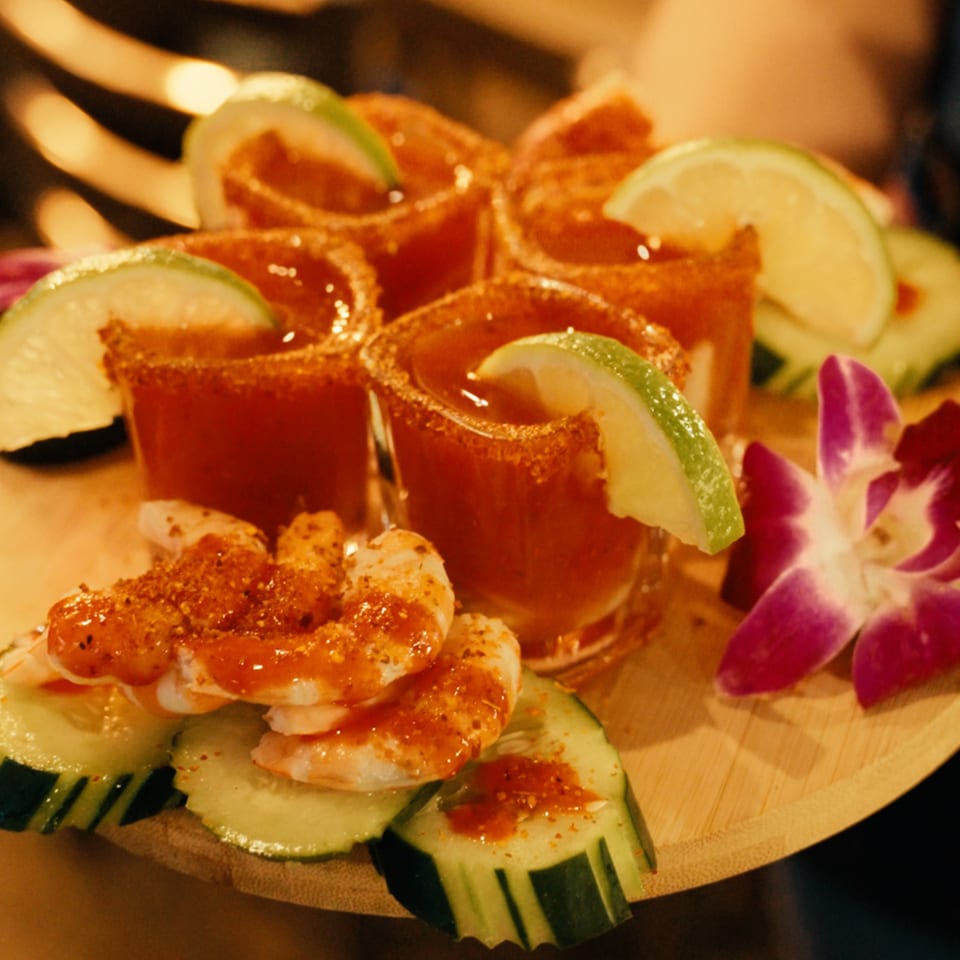 Shrimp and cucumber slices with six shot glasses of red liquid, garnished with lime and a purple orchid.
