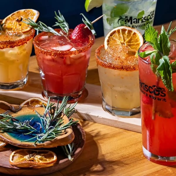Four vibrant cocktails, two red and two yellow, garnished with fruit and herbs on a wooden surface.