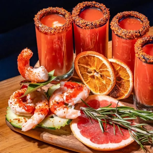 Four spicy red cocktails with shrimp, cucumber, grapefruit, dried orange, and rosemary on a wooden board.