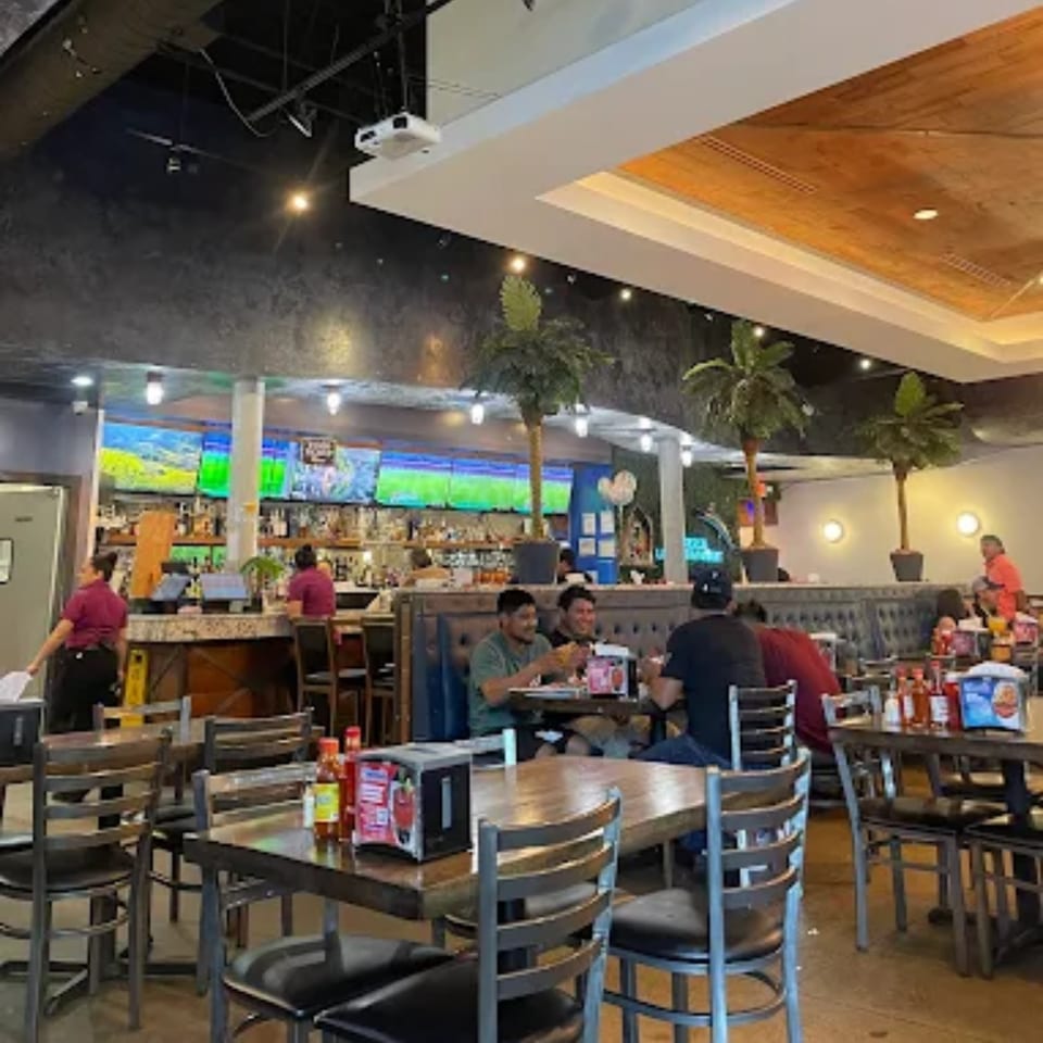 Interior of Mariscos La Riviera Nayarit, showing dining tables, a bar with TVs, and patrons.
