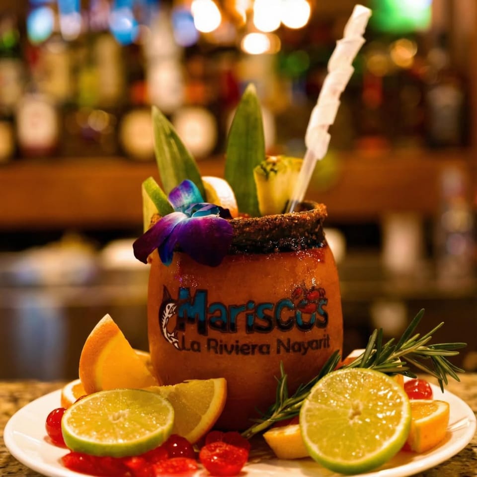A large, garnished cocktail from Mariscos La Riviera Nayarit, with limes, oranges, and cherries on a plate.